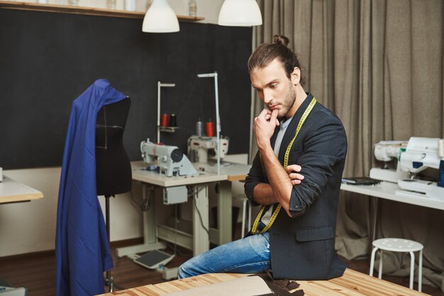 Portret atrakcyjnego dojrzałego skoncentrowanego projektanta mody siedzącego w swoim warsztacie, patrzącego na bok, trzymającego podbródek ręką, myślącego o temacie kolejnej kolekcji ubrań