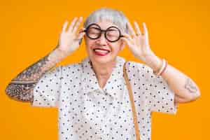 Bezpłatne zdjęcie portret atrakcyjnego funky azjatyckiego tatuażu wesoły siwy starszy starsza starsza kobieta dotyka okularów na białym tle nad żółtym pastelowym kolorem tłaniespodzianka i nad ekscytacją uczuciem