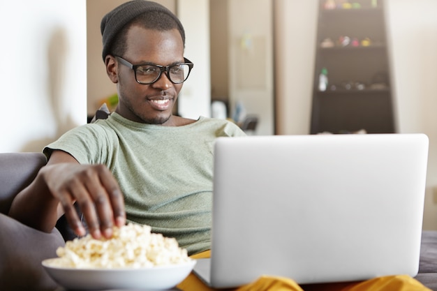 Portret atrakcyjnego młodego samotnego afrykańskiego mężczyzny w okularach odpoczywającego w pomieszczeniu, siedzącego na szarej sofie z laptopem na kolanach, patrzącego z zainteresowaniem na ekran, czytającego e-booka i jedzącego popcorn