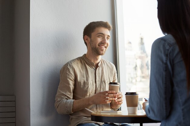 Portret atrakcyjnego, nieogolonego młodego faceta o ciemnych włosach, uśmiechającego się, pijącego kawę i słuchającego opowieści dziewczyn o ciężkim dniu w pracy. Styl życia, koncepcja relacji