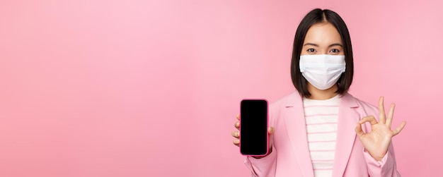 Portret Azjatyckiej Bizneswoman W Medycznej Masce Na Twarz Covid19 Pokazujący Ekran Smartfona I Dobrze Znak Różowy Tło