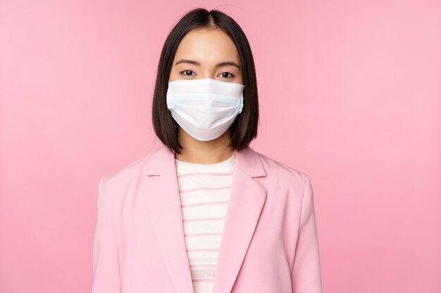 Portret azjatyckiej bizneswoman w medycznej masce na twarz, noszącej koncepcję garnituru pracy biurowej podczas pandemii covid19 stojącej na różowym tle