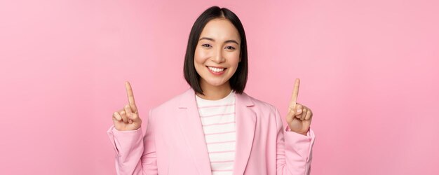 Portret azjatyckiej bizneswoman wskazujący palce w górę i uśmiechający się pokazujący informacje o logo firmy biznesowej na górze stojącej na różowym tle