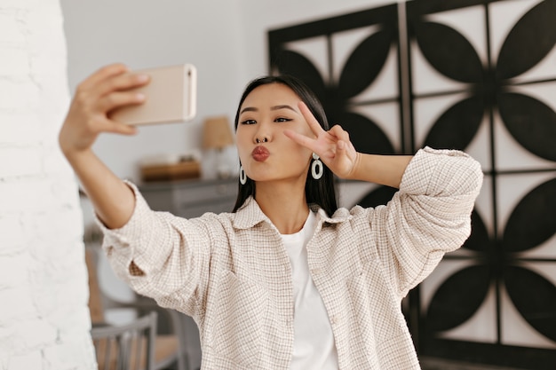 Portret Azjatyckiej Brunetki W Beżowej Kurtce Robi Selfie W Przytulnym Pokoju