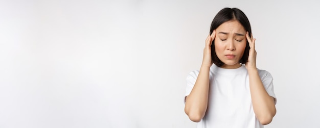 Portret azjatyckiej dziewczyny odczuwającej migrenę głowy lub chorego stojącego w białej koszulce na białym ba