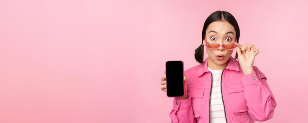 Portret azjatyckiej dziewczyny pokazującej ekran telefonu komórkowego reagującej na zaskoczenie stojącej na różowym tle