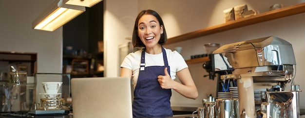 Portret azjatyckiej dziewczyny w fartuchu barista w kawiarni pokazuje kciuki w górę aprobuje lub zaleca smth