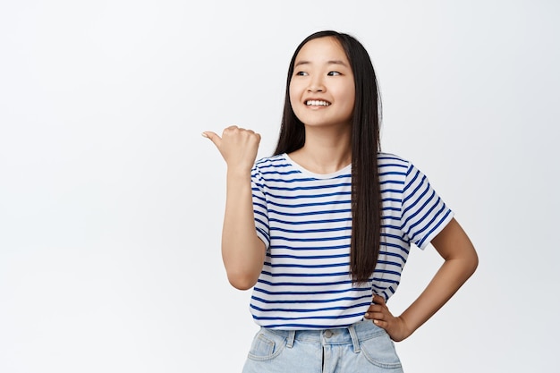 Portret azjatyckiej dziewczyny wskazujący palec w lewo, uśmiechnięty i patrzący na logo firmy, markę na bok, stojącą na białym tle