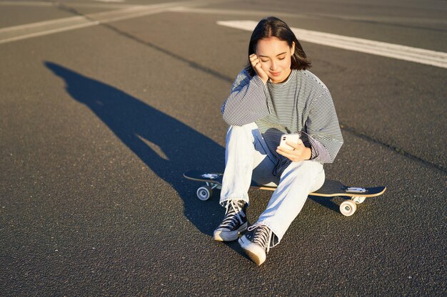 Portret azjatyckiej kobiety siedzącej na deskorolce na łyżwach na swoim krążowniku longboard za pomocą aplikacji na smartfona