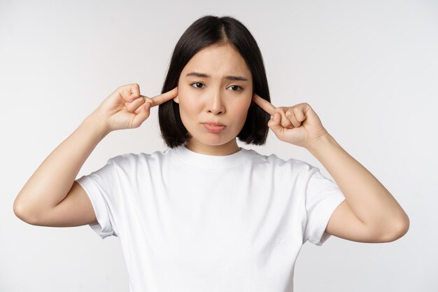 Portret azjatyckiej kobiety zamykającej uszy i odczuwającej dyskomfort z powodu głośnego hałasu denerwującego dźwięku stojącego na białym tle