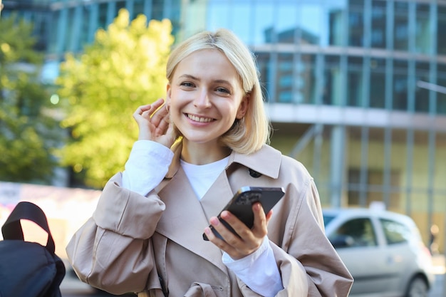 Bezpłatne zdjęcie portret beztroskiej uśmiechniętej kobiety na ulicy trzymającej telefon komórkowy, wkładającej włosy za ucho i patrzącej