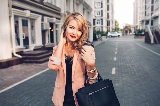Portret blondynka z długimi włosami, chodzenie z kawą w koralowej kurtce na ulicy. Ma winne usta