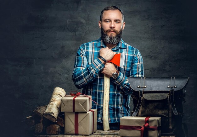 Portret brodatego mężczyzny w średnim wieku trzyma siekierę i stoi w pobliżu stołu z pudełkami na prezenty świąteczne i drewno opałowe na szarym tle.