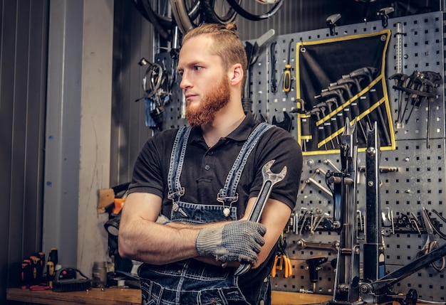 Portret Brodaty mechanik rowerowy ze skrzyżowanymi rękami trzyma klucz do kubka na tle stoiska narzędziowego w warsztacie.