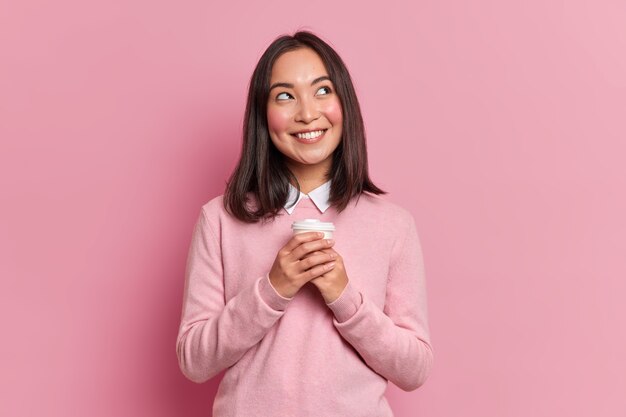 Portret brunetki Azjatki z rozmarzonym wyrazem twarzy uśmiecha się przyjemnie, marząc o kawie na wynos, ubrana w schludny różowy sweter w pozach. Zamyślona modelka dobrze się czuje