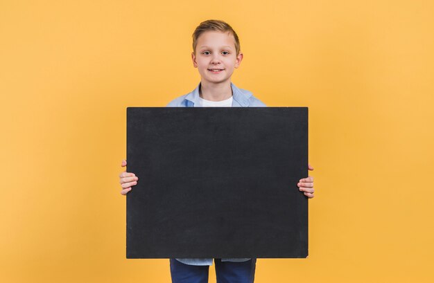 Portret chłopiec trzyma czarną chalkboard pozycję przeciw żółtemu tłu