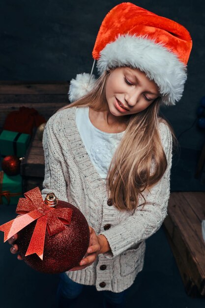 Portret cute teen chłopiec w kapeluszu Świętego Mikołaja, trzymając dużą bombkę w ciemnym pokoju z prezentami.
