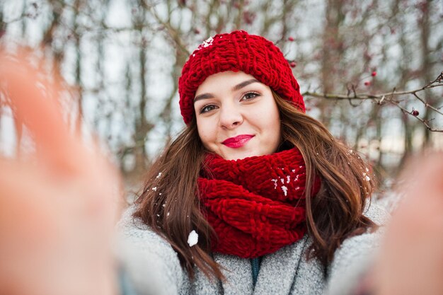 Portret delikatnej dziewczyny w szarym płaszczu, czerwonym kapeluszu i szaliku w pobliżu gałęzi ośnieżonego drzewa trzymającego aparat do selfie