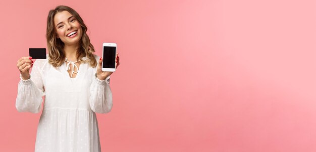Portret delikatnej kobiecej młodej blond dziewczyny w białej sukni, przechyl głowę i uśmiechając się zadowolony rada pobierz aplikację trzymającą telefon komórkowy karta kredytowa pokaż ekran smartfona różowe tło
