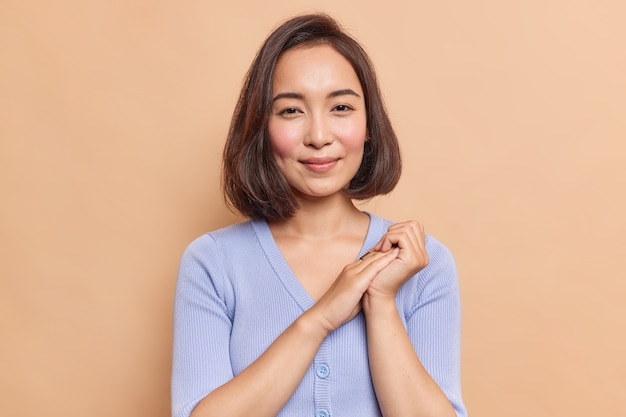 Portret dobrze wyglądająca brunetka młoda azjatycka kobieta cieszy wyrażenie zdrowej skóry naturalne piękno trzyma ręce razem wygląda tajemniczo z przodu ubrana w niebieski sweter pozuje sam w pomieszczeniu