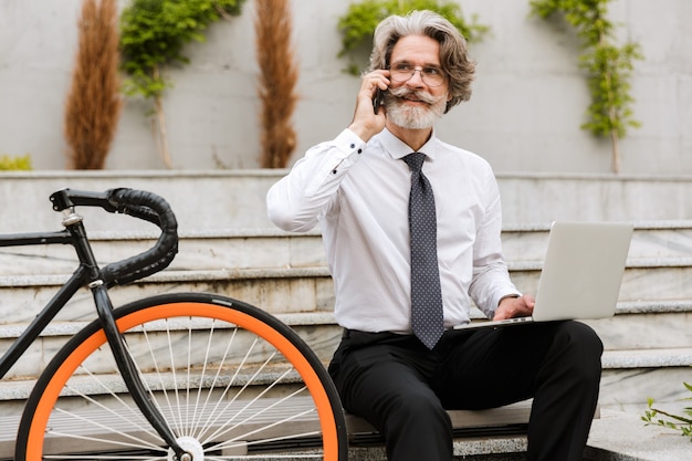Portret dojrzałego, zadowolonego biznesmena w okularach, korzystającego z telefonu komórkowego i laptopa, siedząc na ławce z rowerem na świeżym powietrzu