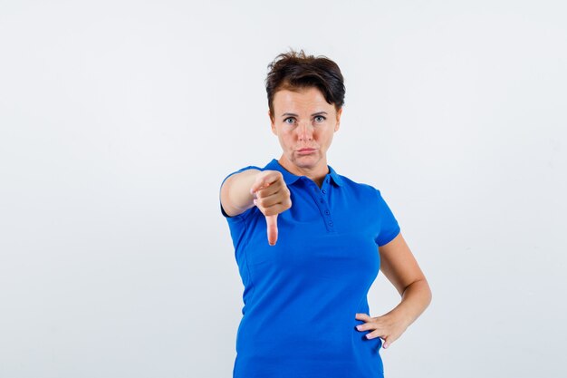 Portret dojrzałej kobiety pokazując kciuk w dół w niebieskiej koszulce i patrząc niezadowolony widok z przodu