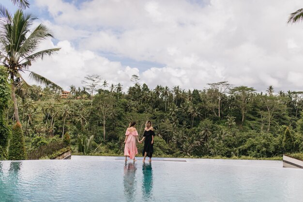 Portret dwóch kobiet pozowanie w pobliżu odkrytego basenu w egzotycznym kurorcie. Zdjęcie wdzięcznych pań w sukienkach stojących na naturze.