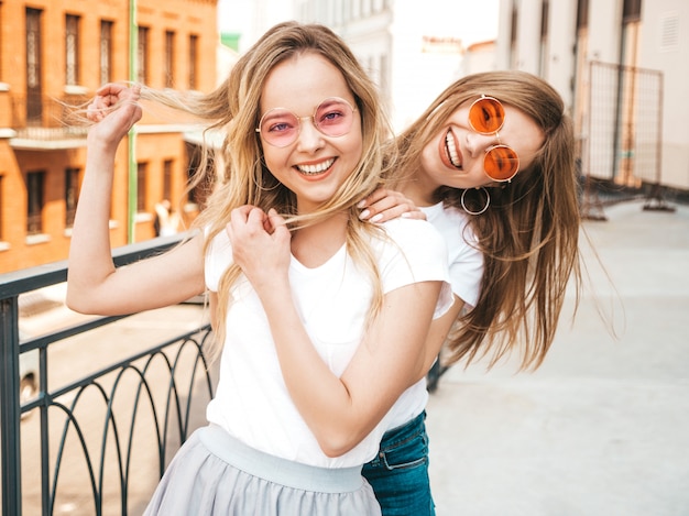 Portret dwóch młodych pięknych blond uśmiechnięte dziewczyny hipster w modne letnie białe ubrania t-shirt. . Pozytywne modele zabawy w okularach przeciwsłonecznych