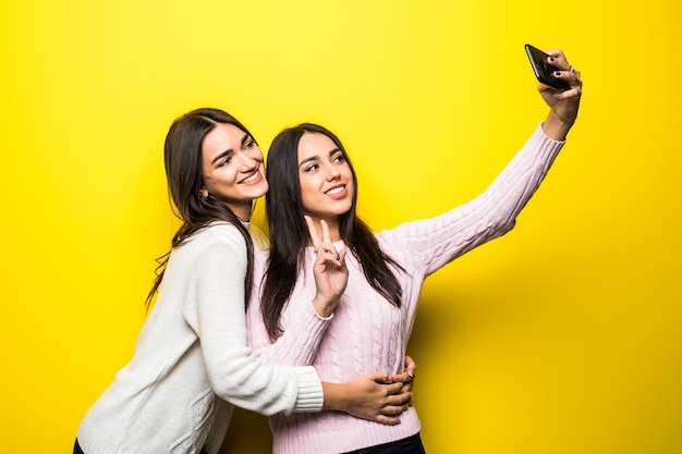 Portret dwóch pięknych dziewczyn ubranych w swetry, stojąc i biorąc selfie na białym tle nad żółtą ścianą