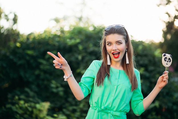 Portret emocjonalnie i piękna brunetka dziewczyna w zielonej sukience, trzymająca cukierka jak panda na patyku i zaskoczony krzykiem. Ładna i stylowa kobieta z czerwonymi ustami, długimi włosami wskazującymi na bok.