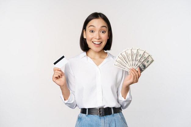 Portret entuzjastycznej azjatyckiej kobiety trzymającej pieniądze w gotówce i karcie kredytowej uśmiechający się zdumiony na białym tle kamery
