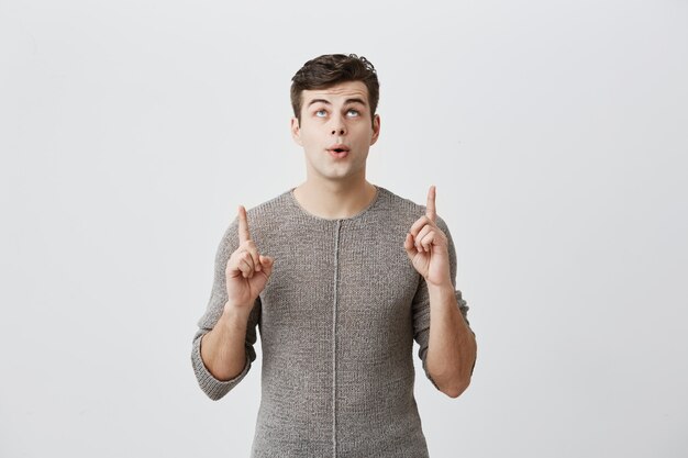 Portret europejski przystojny mężczyzna zaskakuje czymś, ubrany w sweter, wskazując palcem w górę przy pustej ścianie z miejsca kopiowania treści reklamowych lub promocyjnych.