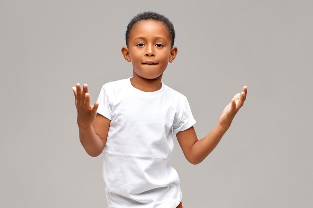 Portret fajnego, uroczego chłopca z Afroamerykanów ubranego w dorywczo białą koszulkę z pewnym wyrazem twarzy pokazującym gest rękami, gryząc dolną wargę. Koncepcja dzieci i stylu życia
