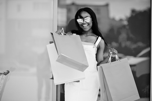 Portret fantastycznej Afroamerykanki w okularach przeciwsłonecznych trzyma wielokolorowe torby na zakupy poza centrum handlowym Czarno-białe zdjęcie