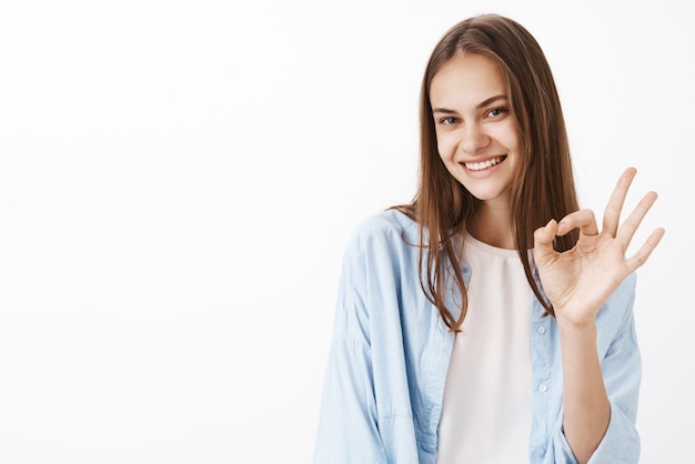 Portret kobiecej szczęśliwej i pewnej siebie stylowej brunetki w niebieskiej modnej bluzce na białej koszulce pokazującej gest w porządku lub w porządku i uśmiechającej się z pewnym spojrzeniem