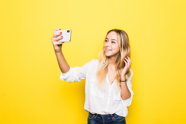 Portret kobiety biorą selfie, trzymając w ręku inteligentny telefon, fotografowanie selfie na białym tle na żółtej ścianie