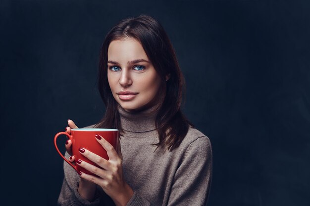 Portret kobiety brunetka ubrana w brązową kurtkę z długą szyją trzyma filiżankę czerwonej kawy.