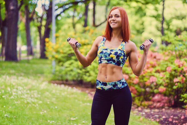 Portret kobiety fitness rude trzyma hantle w parku na świeżym powietrzu.