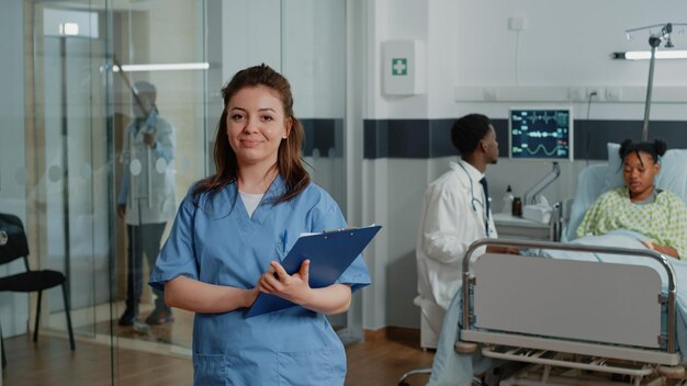 Portret kobiety pracującej jako pielęgniarka trzymająca papiery kontrolne, stojąc na oddziale szpitalnym. Asystent medyczny z dokumentami patrzący w kamerę i uśmiechnięty, udzielający pomocy specjaliście.