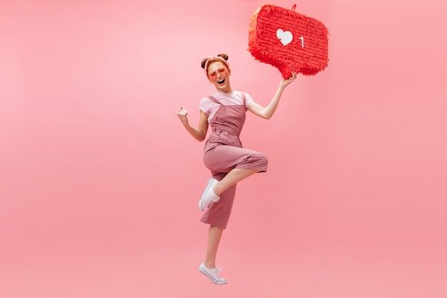 Bezpłatne zdjęcie portret kobiety rude w różowe okulary i kombinezon, raduje się ze zwycięstwa i skacze na na białym tle.