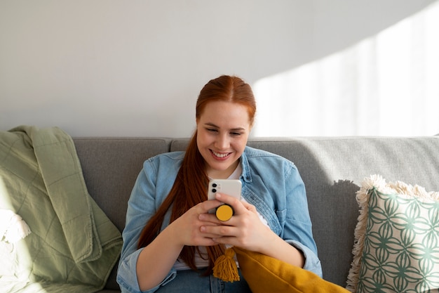 Portret kobiety używającej smartfona w domu na kanapie, trzymając ją z gniazda pop