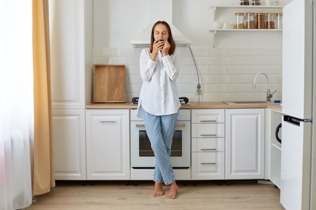 Portret kobiety w białej koszuli i dżinsach stojącej, pachnącej filiżanką kawy w kuchni, pachnącej aromatycznym napojem rano, pozując w kuchni.