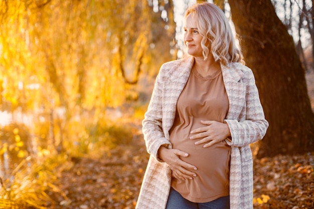 Portret kobiety w ciąży w średnim wieku na zewnątrz w parku Kobieta w średnim wieku spodziewa się dziecka w wieku ciąży Blond kobieta ubrana w brązowy sweter i beżowy płaszcz