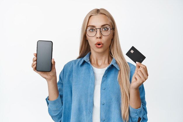 Portret kobiety w okularach reagującej na zaskoczenie pokazujący ekran telefonu komórkowego z kartą kredytową, wpatrującą się w kamerę białe tło