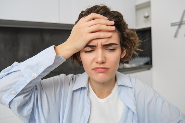 Portret kobiety z bólem głowy siedzi w kuchni, dotyka głowy i krzywi się, ma bolesną migrenę