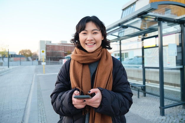 Portret koreańskiej kobiety w kurtce zimowej stojącej ze smartfonem czekającej na autobus na przystanku patrząc a