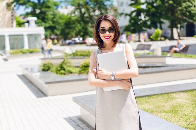 Portret ładna brunetka dziewczyna w szarej sukience stojącej w parku w mieście. Trzyma laptopa i uśmiecha się do kamery.