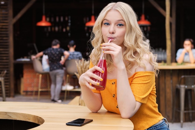 Portret ładnej blond dziewczyny patrzącej w aparacie podczas picia lemoniady na dziedzińcu kawiarni