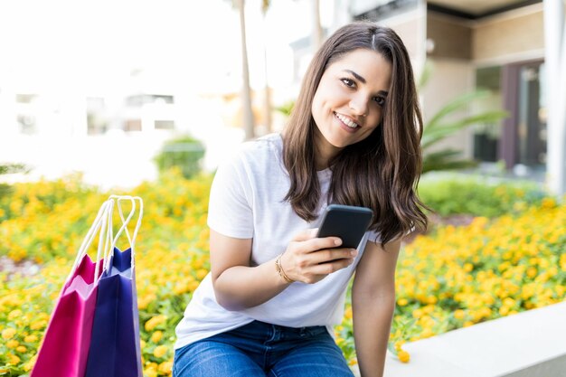 Portret ładnej kobiety trzymającej smartfon siedzący przy torbach na zewnątrz centrum handlowego