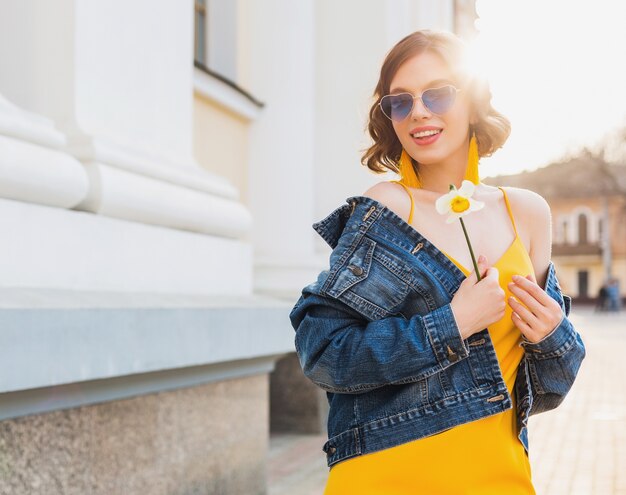 Portret ładnej kobiety w okularach przeciwsłonecznych w kształcie serca, trzymającej kwiat przed słońcem, słoneczny letni dzień, stylowa odzież, trend w modzie, niebieska kurtka jeansowa, żółta sukienka, eleganckie kolczyki boho hipster